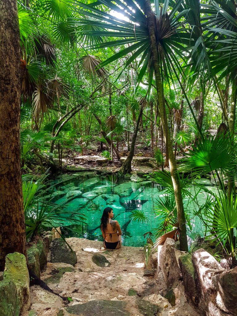 a girl sits on the edge of a turquoise cenote surrounded by lush jungle foliage