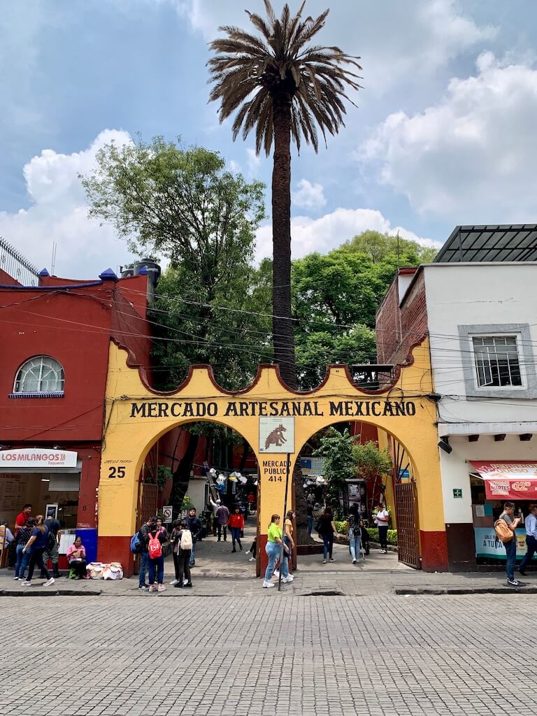 Looking across a cobblestone street to the yellow arches that form the entrance to the Mercado Artesanal Mexicano in Coyoacan, Mexico City