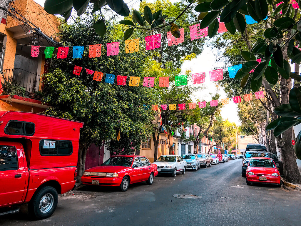 papel picado is strung across a street in Coyoacan, Mexico. Red cars and leafy green trees line the sides of the street.