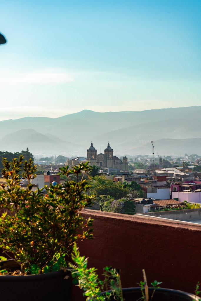 A view from a rooftop in Oaxaca, overlooking a scenic landscape with the Santo Domingo de Guzmán church's domes rising against a backdrop of hazy blue mountains, encapsulating the tranquil beauty found in Oaxaca.