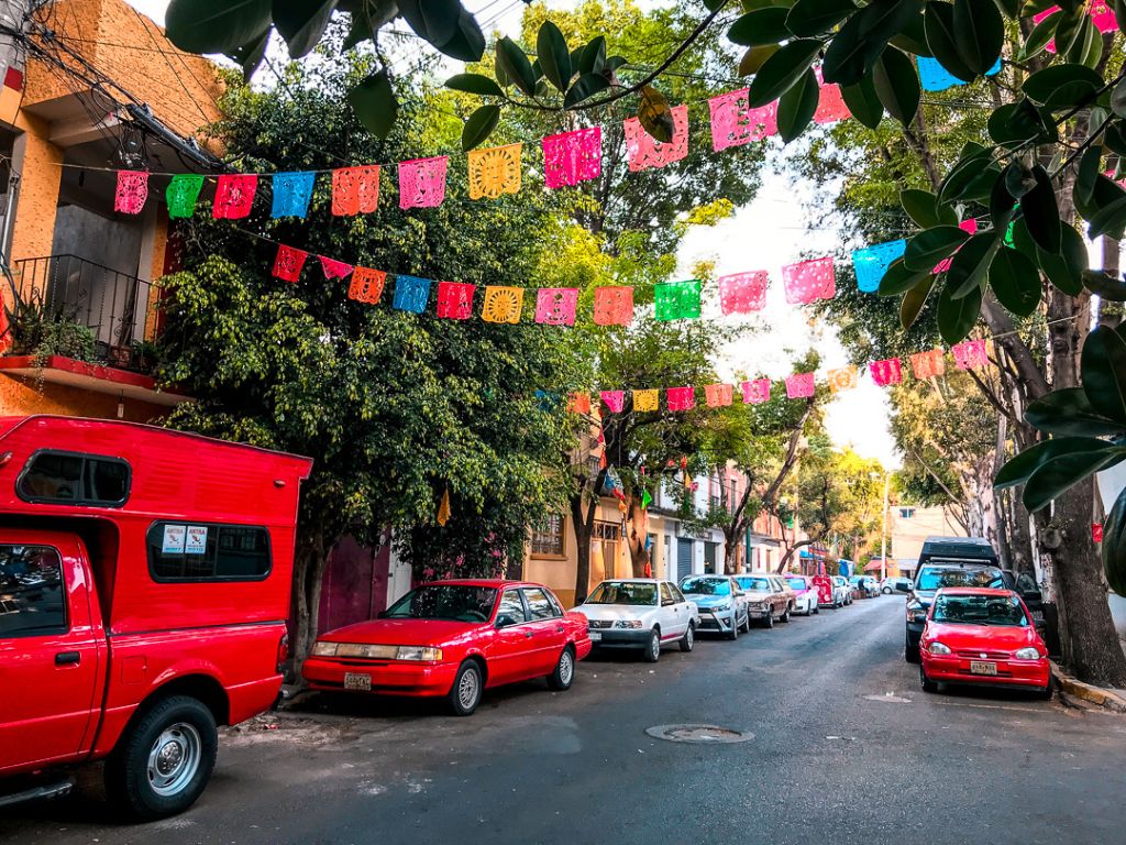 a street in Coyoacan, Mexico City is lined by red cars and trees. Papel picado stretches across the street overhead