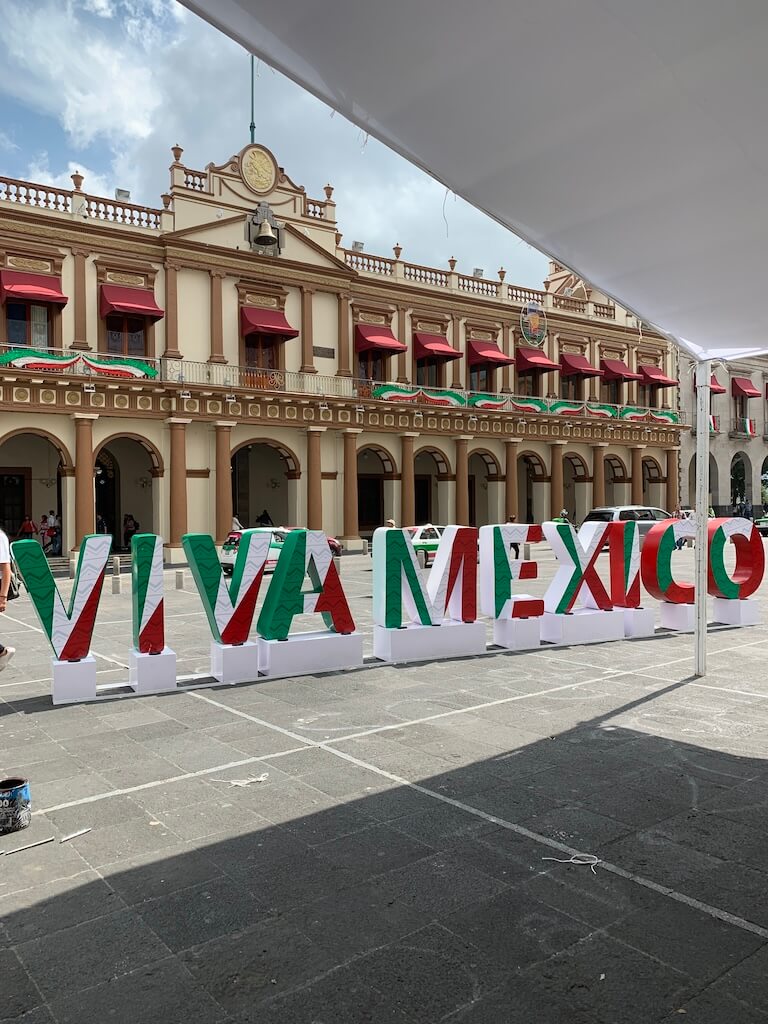 government building in Xalapa, Mexico adorned with red, white, and green ribbons, with a large Viva Mexico sculpture in the foreground
