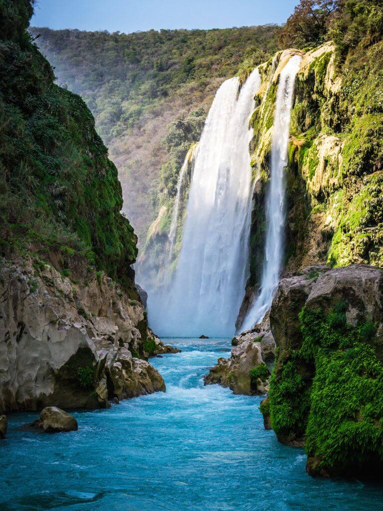 looking down a river with crystalline blue water, at a towering waterfall coming down from lush green hills