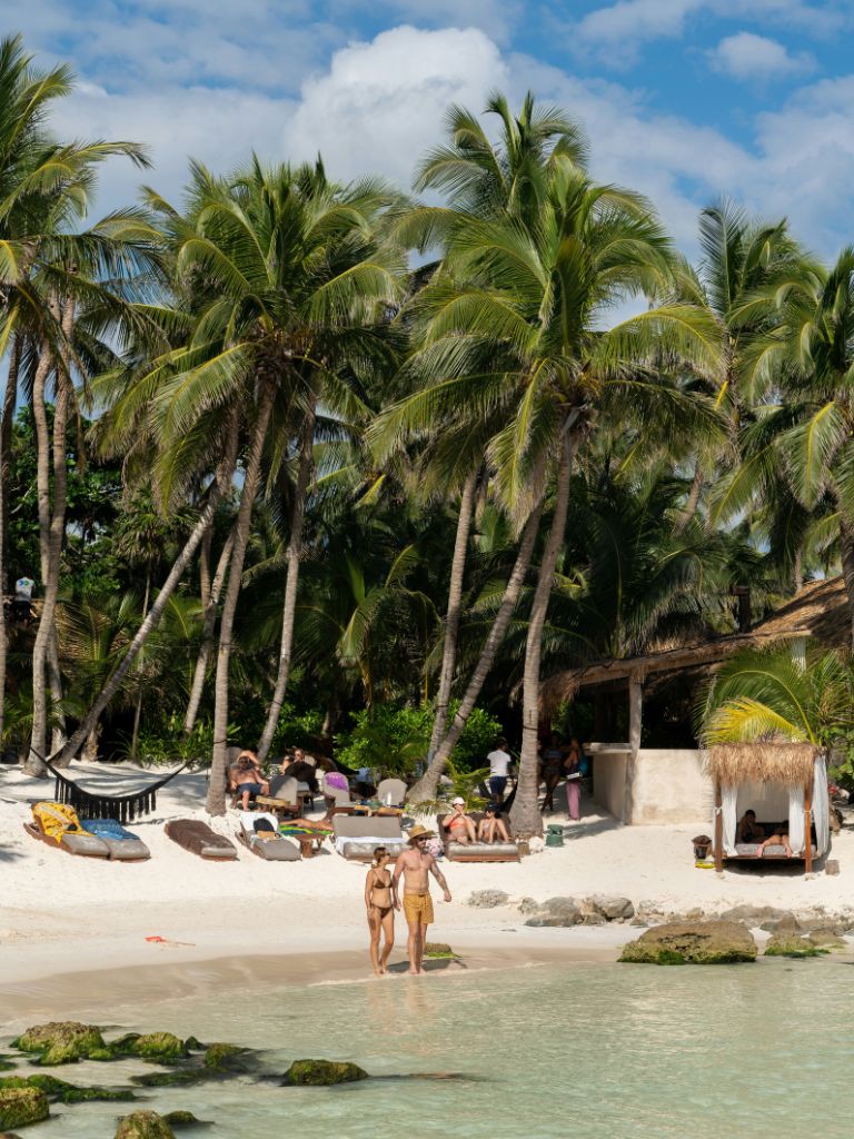 a man and woman in swimsuits wade into the turquoise water at a beach in tulum, other travelers lay on sun loungers in the distance