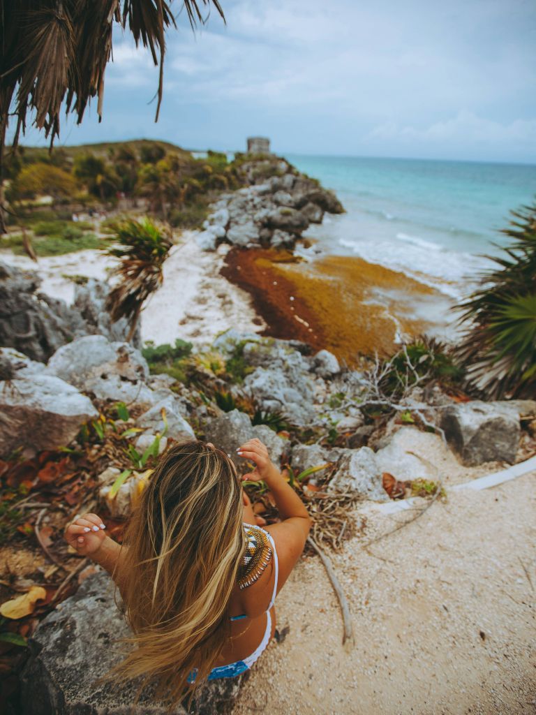 a woman sits on a sandy bluff looking over a whitesand beach with the tulum ruins in the distance